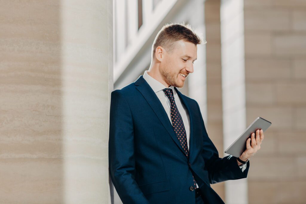 Sideways shot of elegant businessman in formal suit, holds digital tablet in hands and reads business news on internet website, poses in office, connected to wireless wifi. Modern technologies concept